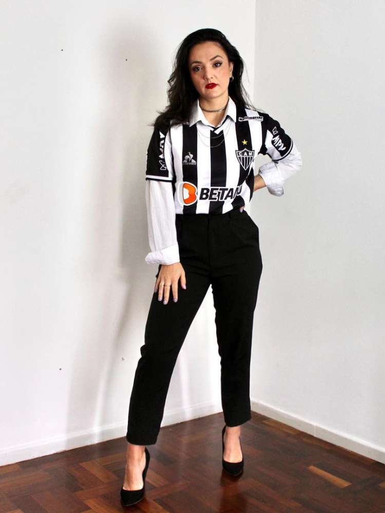 Mulher de pele clara e cabelo escuro usando camisa branca embaixo de camisa do Atlético Mineiro e calça preta curta
