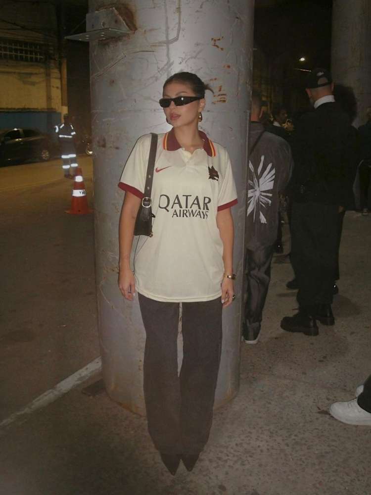 Malu Camargo usando óculos de sol, camisa da Roma, bolsa preta e calça escura