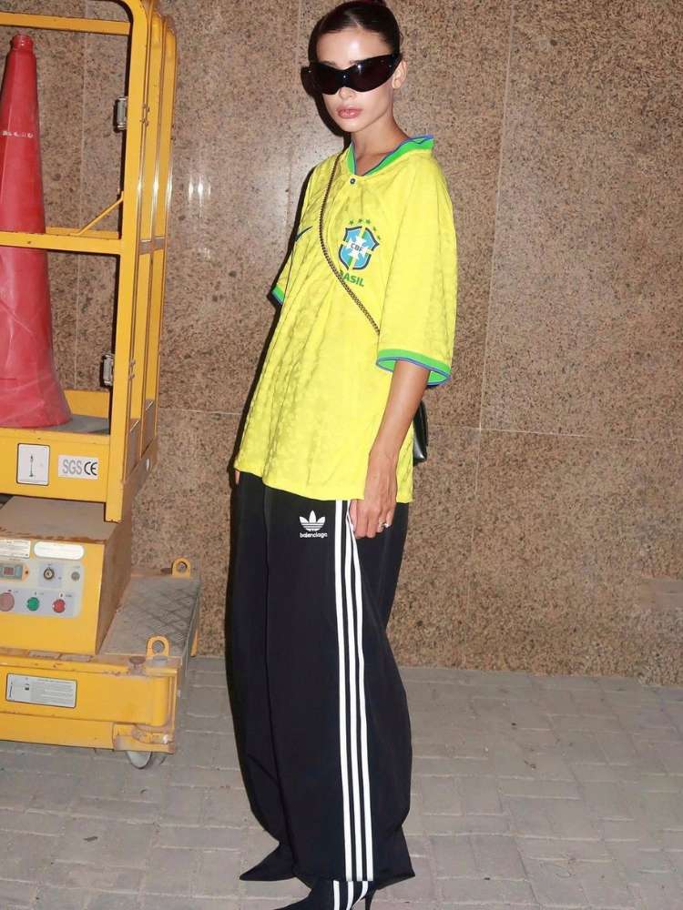 Livia Nunes com camisa amarela da Seleção Brasileira, óculos de sol, calça larga preta da Adidas e salto preto.