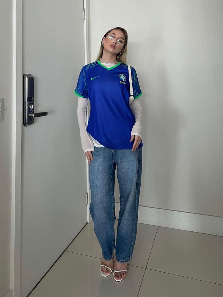 Malu Borges usando camisa azul da Seleção Brasileira sobre blusa de manga comprida branca, calça jeans e bolsa branca