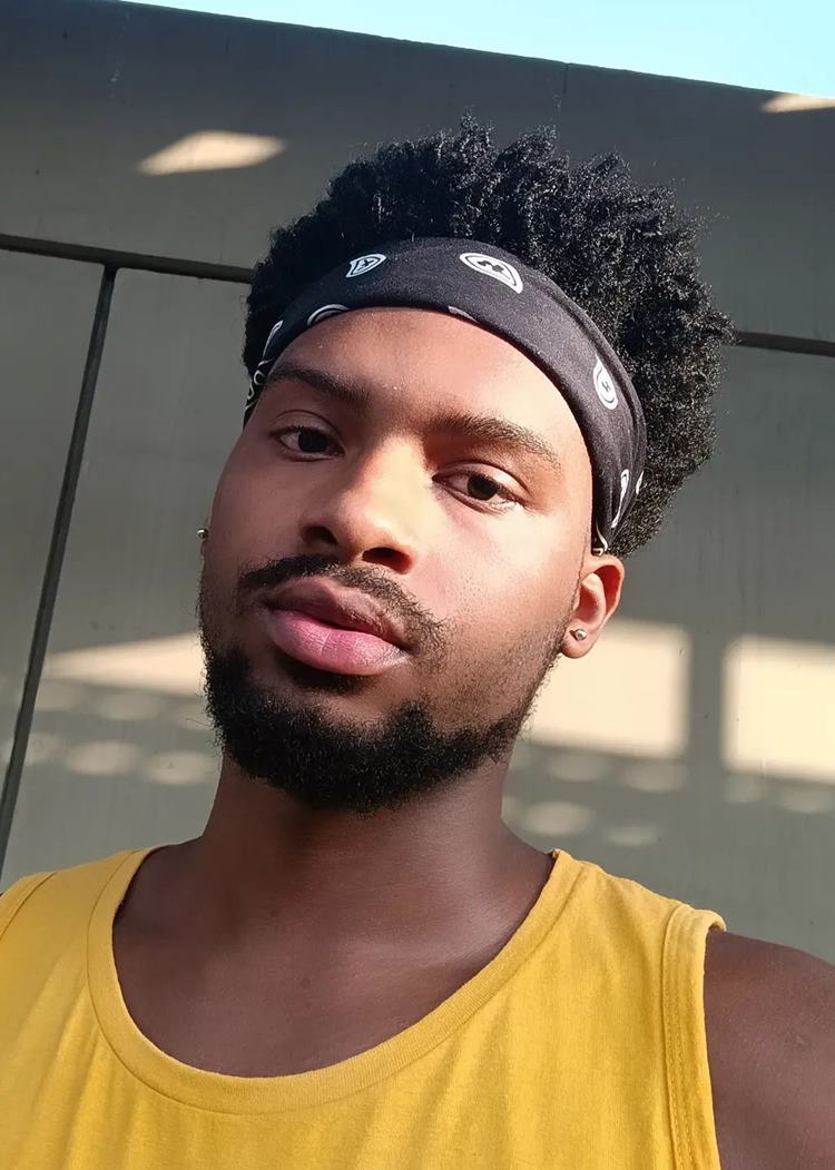 Homem com pelo do rosto estilo Chinstrap moderno. Ele tem o cabelo lcacheado, usa bandana, regata amarela em fundo de muro