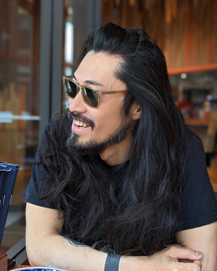 Homem com pelo do rosto estilo Chinstrap moderno. Ele tem o cabelo longo, está com camiseta preta, de óculos de sol, sentado em cadeira de uma cafeteria possivelmente