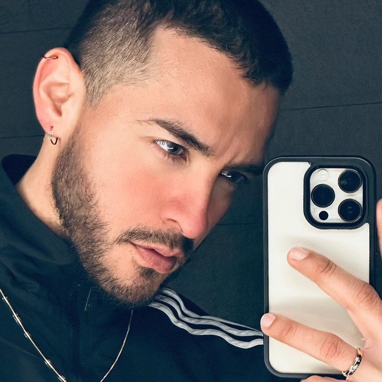 Homem com pelo do rosto estilo Chinstrap moderno. Ele tem o cabelo preto, está com roupa de frio e tirando selfie no espelho