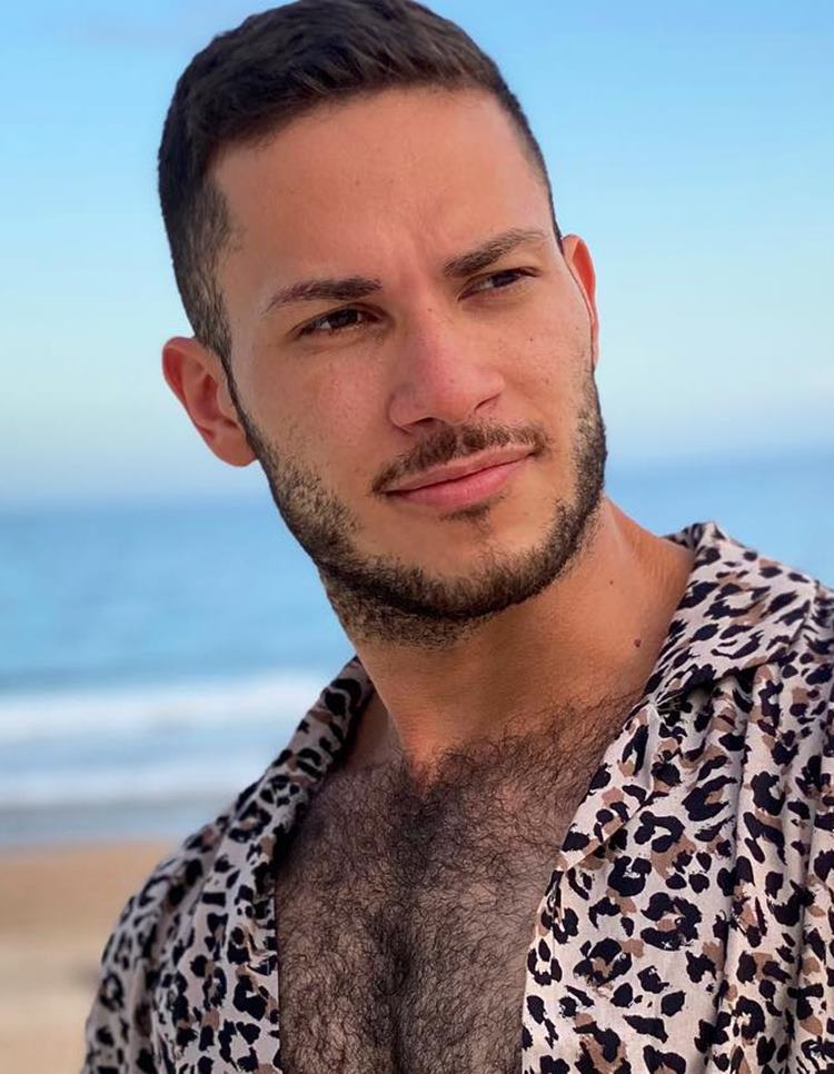 Homem com pelo do rosto estilo Chinstrap moderno. Ele tem o cabelo preto, usa camisa com estampa de onça aberta e está na praia