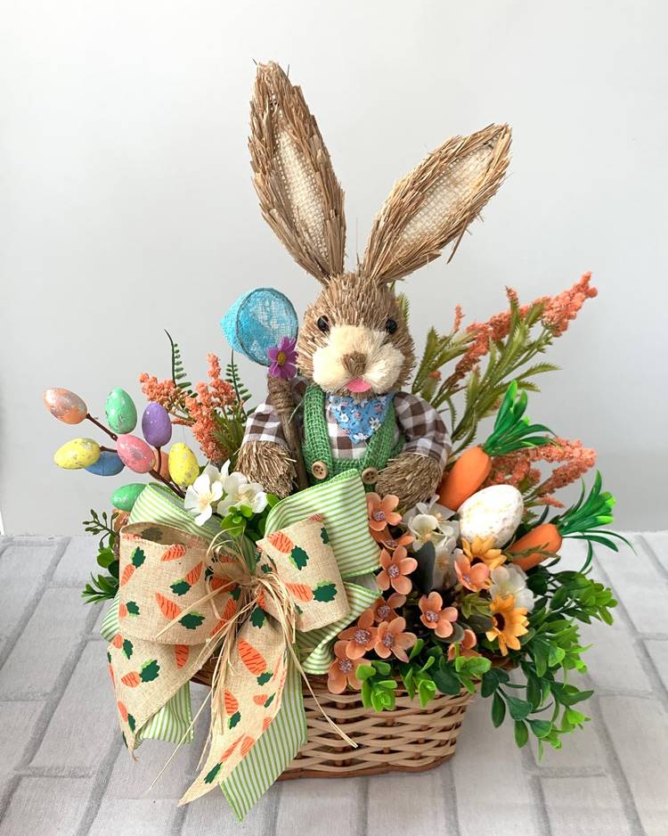 Cesta contendo um coelhinho artesanal feito de palha e tecido. A cesta está repleta de várias flores e decorações.