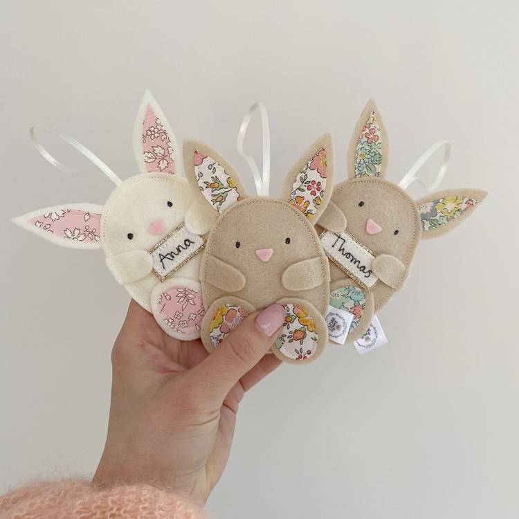 Mão segurando três coelhinhos de feltro, personalizados com nomes, funcionando como uma espécie de chaveiro.