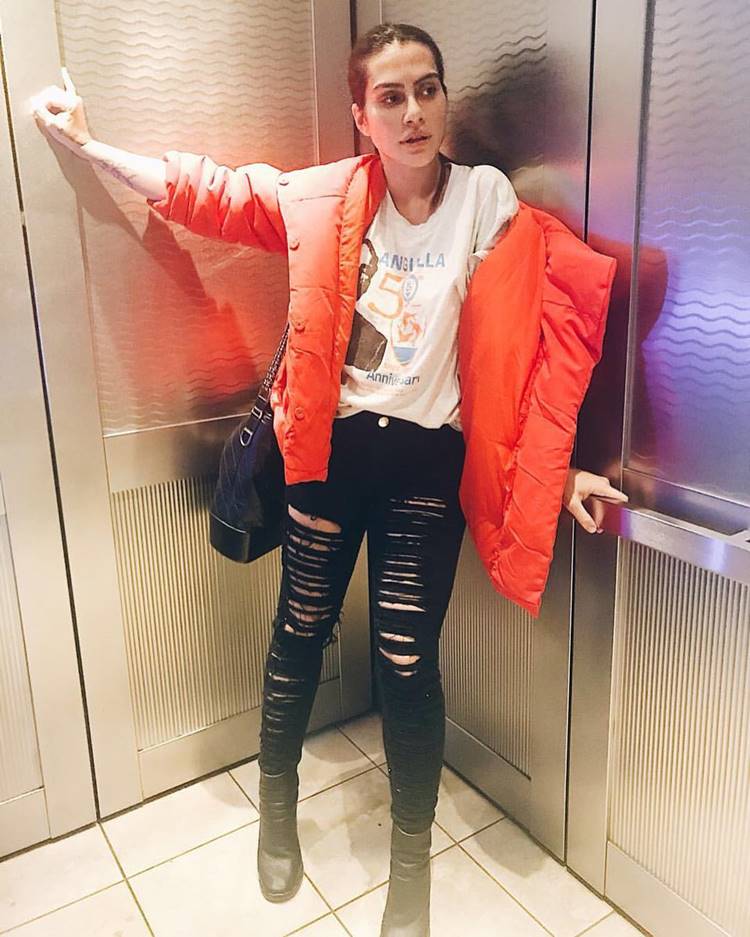 Elevador Fashion: Presumivelmente dentro de um elevador, ela se destaca com uma camisa estampada, calça jeans rasgada, botas, várias bolsas e uma jaqueta laranja puffer, um mix de casualidade e tendência.

