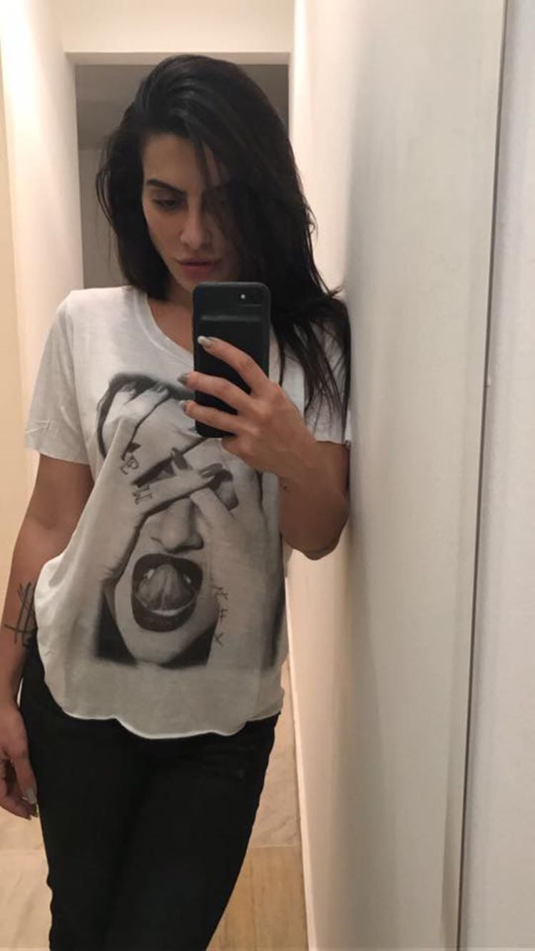 Autoretrato Casual: Em outra cena do passado, ela aparece vestindo uma camisa estampada e calça preta, encostada em uma parede enquanto captura uma selfie no espelho, um momento casual e íntimo.

