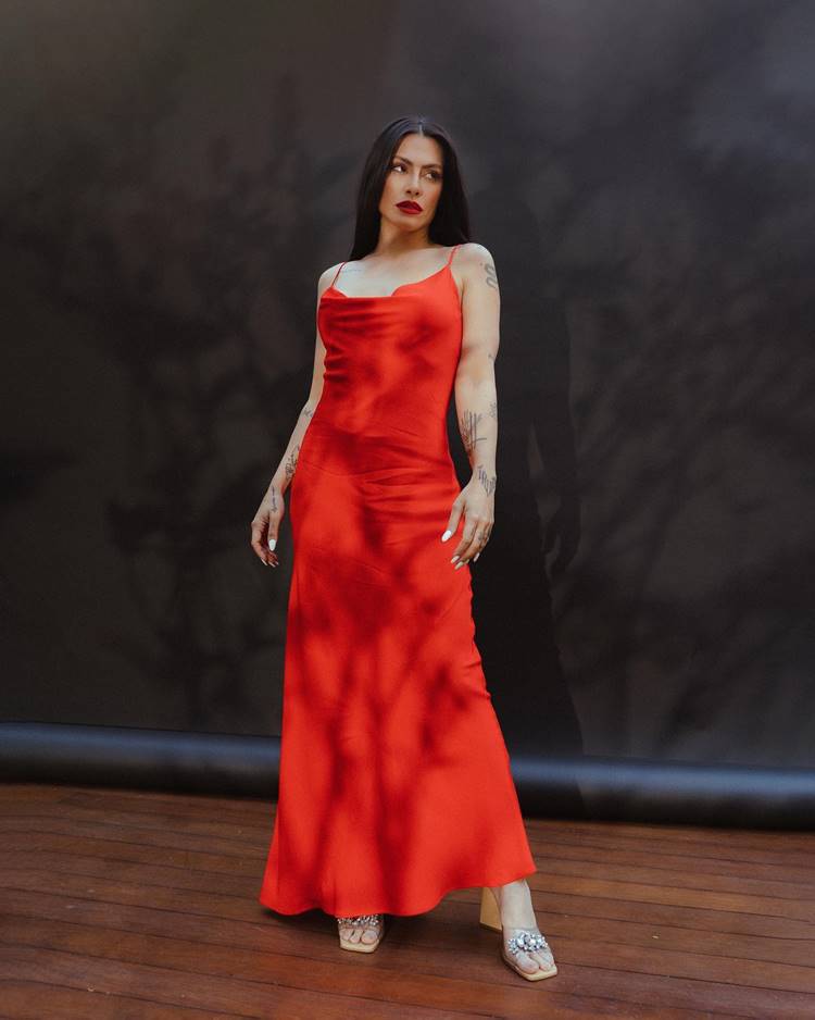 Seda e Sedução: A atriz usa um vestido longo vermelho de seda, sandália de salto e maquiagem com batom vermelho, diante de um fundo preto, uma representação clássica de glamour e sensualidade.

