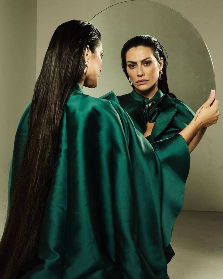 Reflexão em Verde: Segurando um espelho, a atriz veste uma roupa de seda verde, uma imagem introspectiva que também celebra a beleza e a simplicidade.

