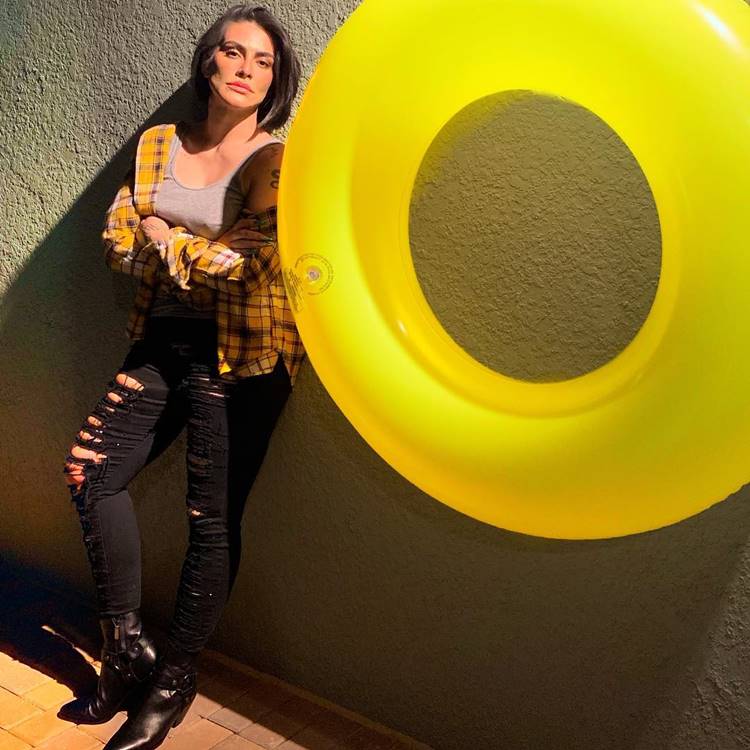 Xadrez e Jeans Rasgado: Com uma regata cinza, camisa xadrez amarela e calça jeans preta rasgada, complementada por botas de salto, ela se encosta em uma parede adornada com uma boia amarela, um estilo rocker descomplicado.

