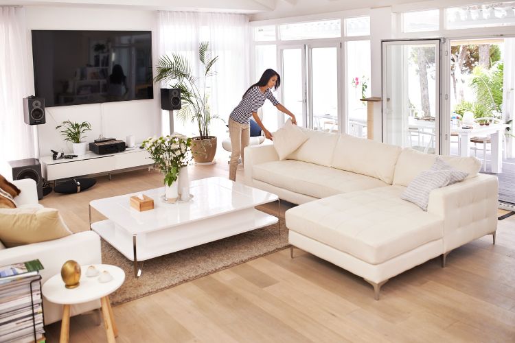 Mulher de pele clara arrumando almofada de sofá bege em sala de estar com chão de madeira, TV e janelas grandes