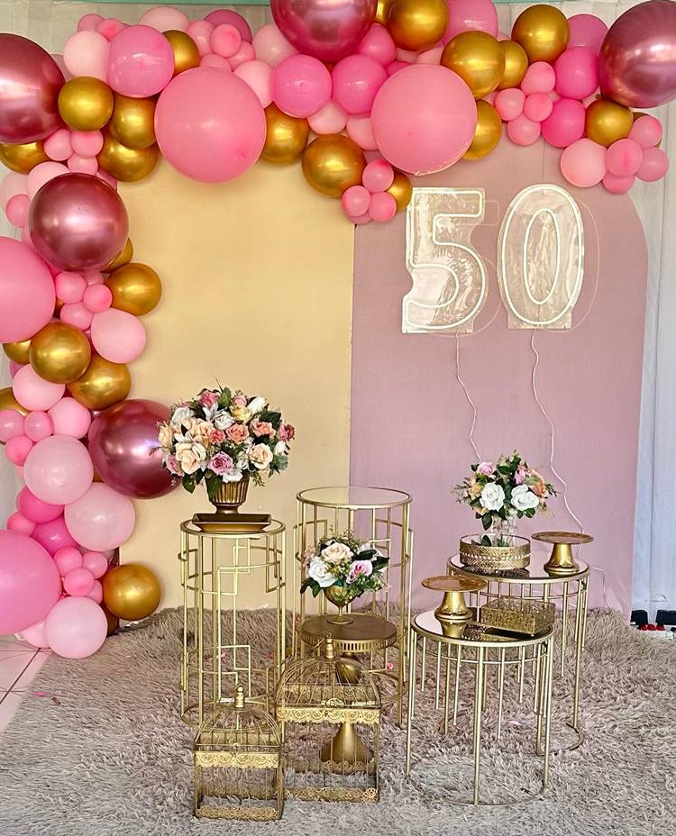 Placa de "50 anos" ao fundo de decoração de festa rosa e dourada