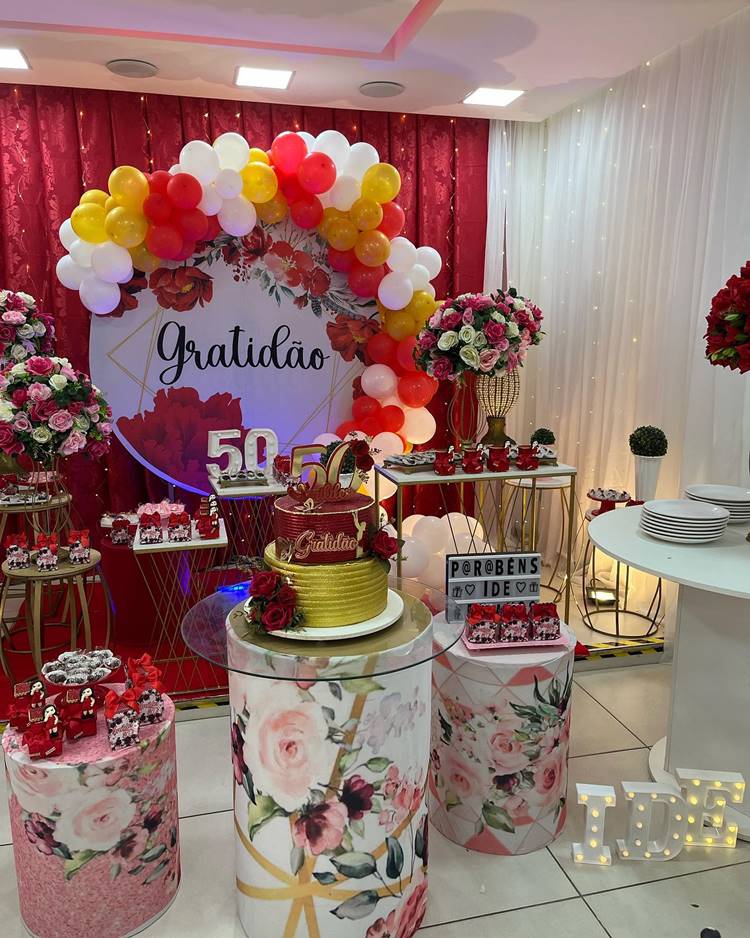 Esta festa de 50 anos brilha em tons de vermelho, branco e rosa, com uma mensagem de gratidão permeando o tema floral. O cenário é composto por um bolo elegante, doces finamente decorados, rosas frescas e plaquinhas temáticas, celebrando a ocasião com estilo e cor.