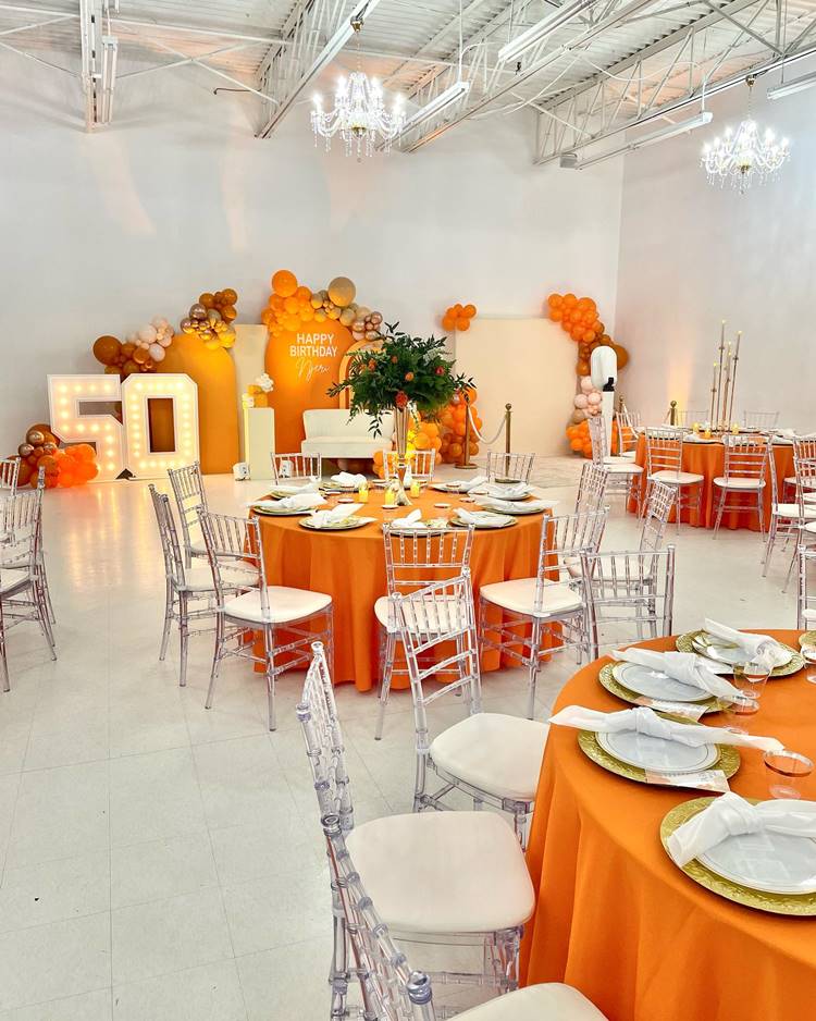 O salão está arranjado com mesas redondas adornadas com centros de mesa florais e velas, criando um ambiente acolhedor. Ao fundo, destaca-se um vibrante painel laranja com balões e um espaço dedicado para fotos, iluminado por uma placa com o número 50, celebrando o marco de meio século.