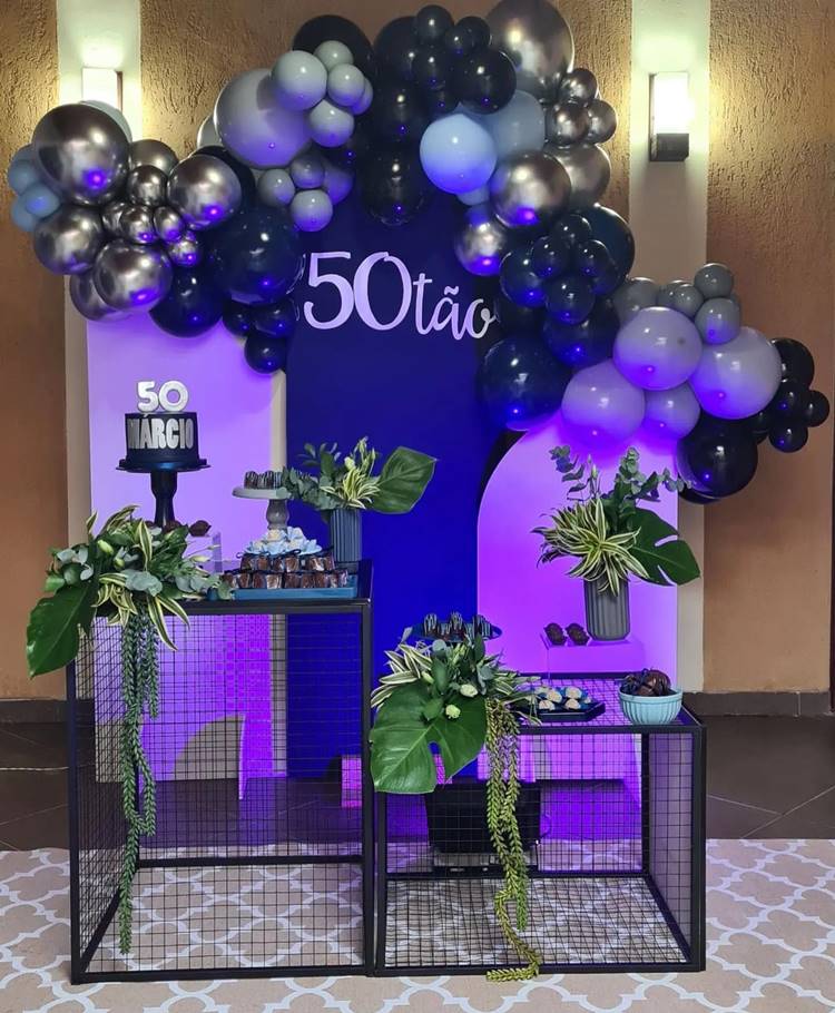 Esta festa, dedicada a um homem que alcança o marco dos 50 anos, apresenta uma paleta de azul e branco, com balões em tons de cinza e preto. Detalhes em verde das folhas de plantas e um bolo pequeno marcado com o número 50 sob uma iluminação roxa, criam um ambiente sofisticado para a celebração.