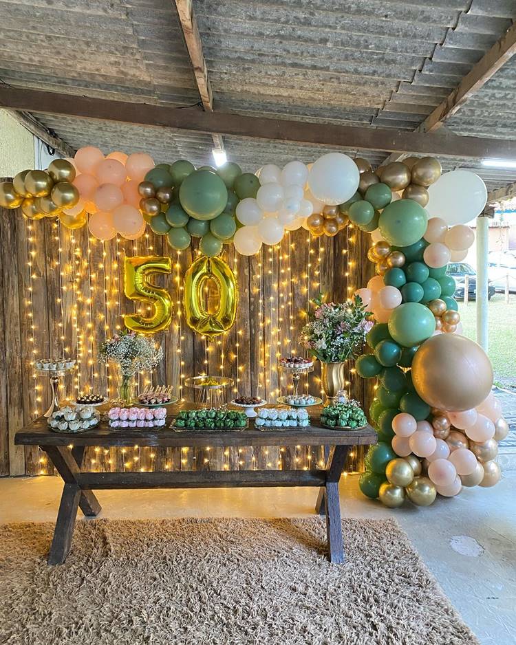 Com um painel de madeira ripada, balões em tons de verde, dourado e branco, e um balão dourado com o número 50, esta decoração rústica celebra o aniversário com um toque natural. A mesa de madeira, colocada sobre um tapete estiloso, é decorada com doces e flores, aguardando apenas o bolo para completar a celebração.