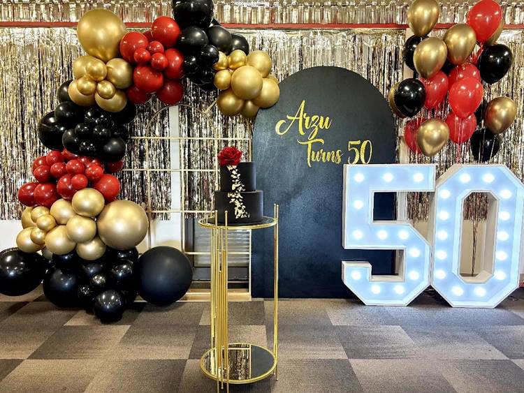 Combinação de dourado, vermelho e preto, com balões, bolo preto e dourado, e uma grande placa iluminada de "50 anos".