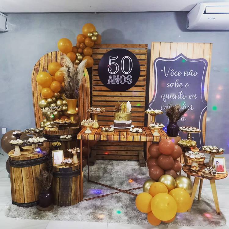 Tema inspirado em madeira com cores marrom, dourado e preto. Destaque para a placa com "50 anos" e a frase emocionante sobre a jornada até aqui. A mesa exibe doces, bolo e fotos da pessoa, celebrando sua história de vida.