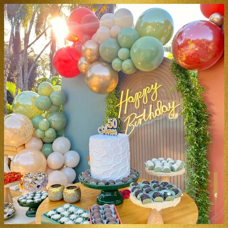  Durante o dia, com balões em vermelho, verde, bege e dourado, um bolo alto e docinhos variados.