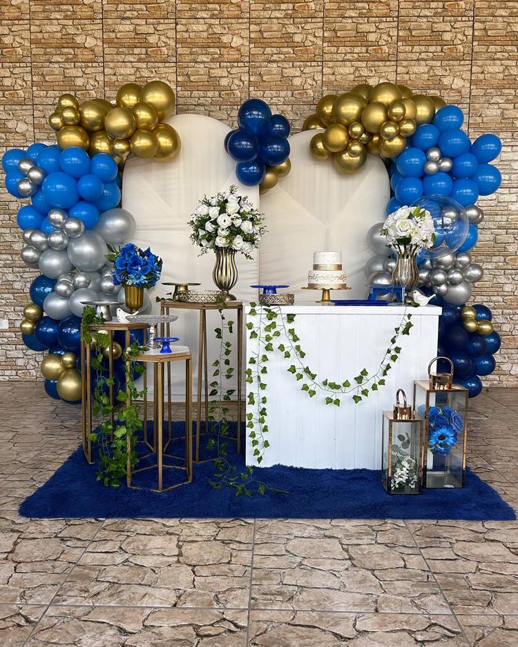 Em um salão que parece pertencer a uma residência, a decoração de 50 anos combina azul, dourado e branco, com flores em vasos e um bolo elegante, complementados por um painel adornado com folhagem de jiboia, celebrando meio século de vida com elegância e serenidade.