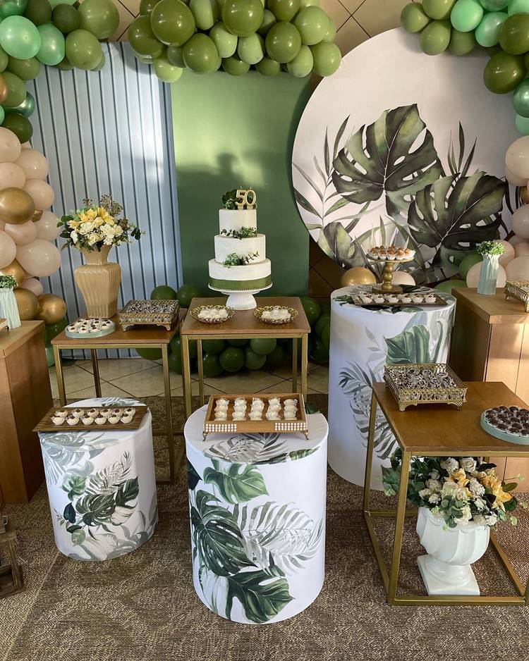 A decoração desta festa de 50 anos é inspirada na natureza, com um foco na folha de costela de Adão, complementada por tons de verde, dourado e branco. O ambiente é enriquecido com doces temáticos, um bolo decorado, flores e balões, criando uma atmosfera harmoniosa e elegante.
