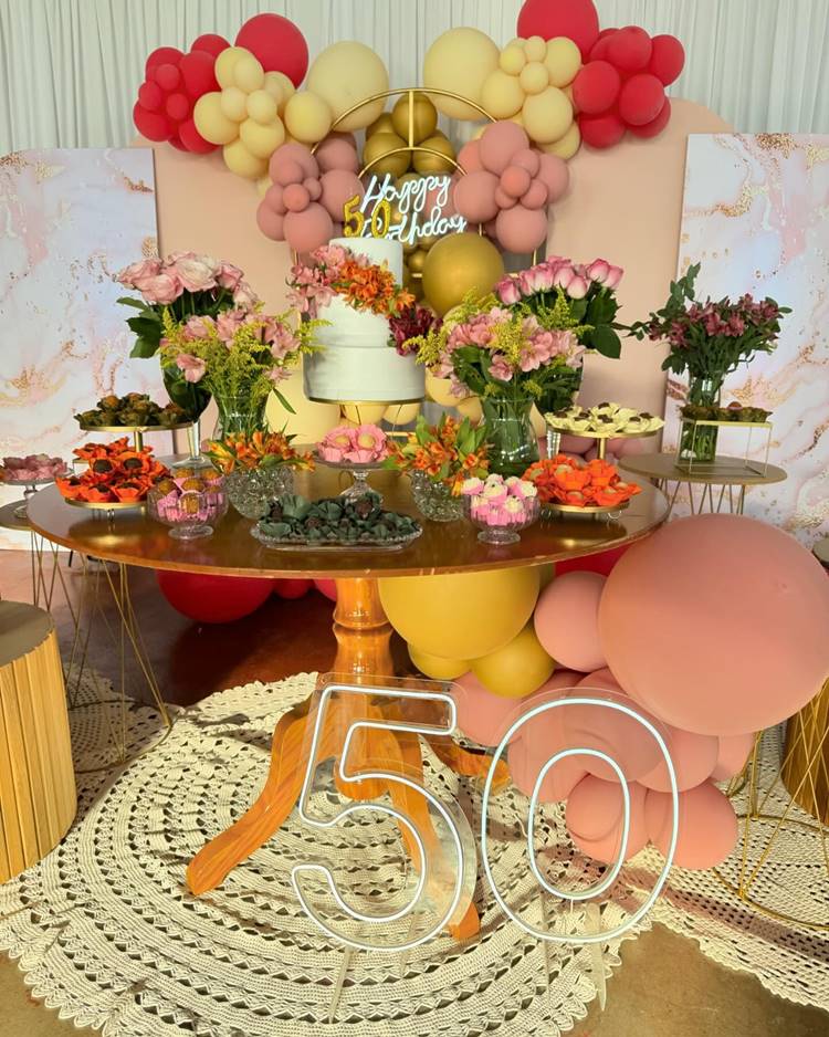 Um painel repleto de flores coloridas serve como pano de fundo para esta festa de 50 anos, que também conta com balões coloridos, um bolo decorativo e uma mesa de madeira ornamentada. Abaixo da mesa, tapetes de crochê e uma plaquinha moderna e transparente comemoram os 50 anos de forma vibrante e alegre.