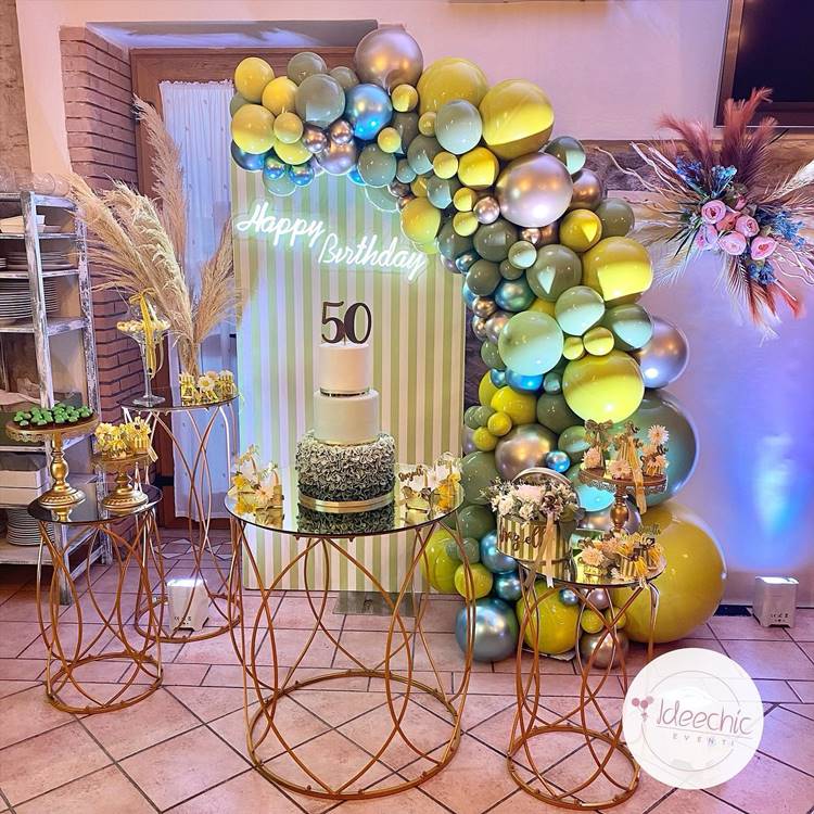 Com uma decoração que utiliza balões em diversos tons de verde, esta festa de 50 anos não segue um tema específico, mas encanta com sua simplicidade. A mesa de metal com tampo de vidro exibe um bolo de três andares, coroado com uma plaquinha de 50, cercado por decorações florais e de trigo, celebrando a ocasião de forma singela mas memorável.