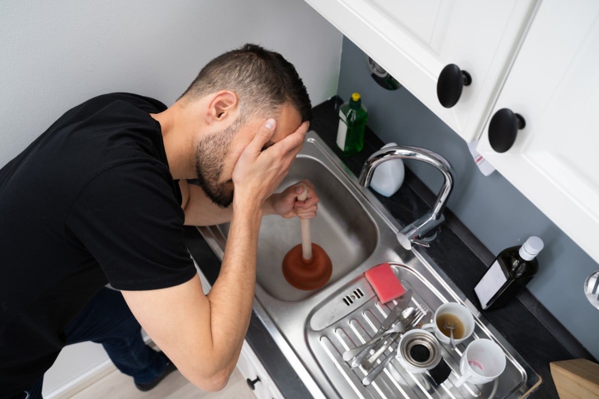 Homem de pele branca usando camiseta preta e com a mão no rosto enquanto segura desentupidor na pia da cozinha com as louças ao lado