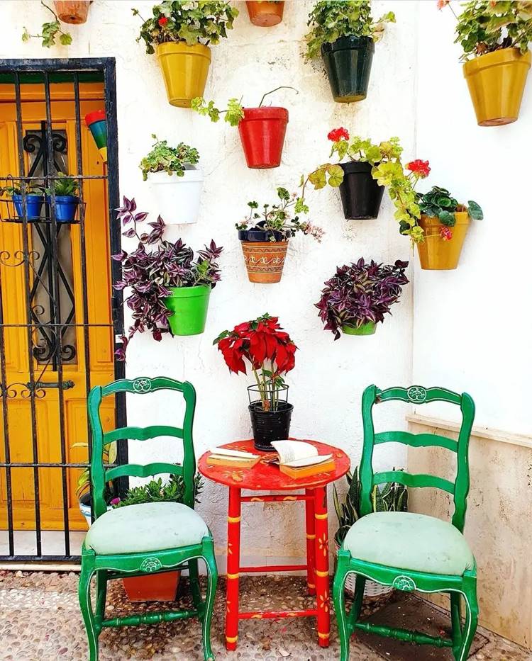 Vasos coloridos com plantas pendurados em parede de área externa, com porta e uma mesa com 2 cadeiras