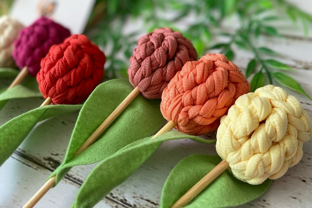 Buquê artesanal com flores feitas de tecido trançado em tons coloridos, incluindo vermelho, laranja e amarelo, sobre hastes de madeira com folhas verdes de feltro. Fundo desfocado com detalhes naturais, ideal para decoração ou lembrancinhas.