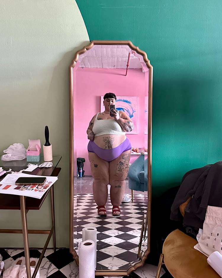 Mulher com lipedema usando croped e shorts curto roxo, em selfie no espelho de quarto