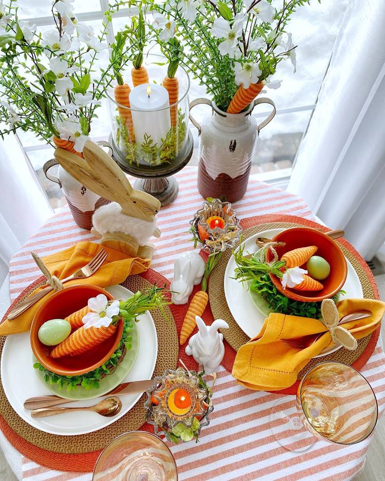 Mesa posta de Páscoa com itens de cor laranja bege marrom e branco a dois lugares na mesa redonda pratos sobrepostos talheres taças e jarros com arranjos