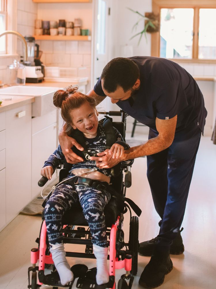 Pai na cozinha fechando cinto da cadeira de rodas da filha, que possui deficiência 