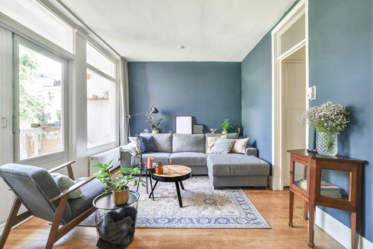 Sala com grande janela, parede azul, tapete com estampa semelhante a azulejo, chão de madeira e sofá azul
