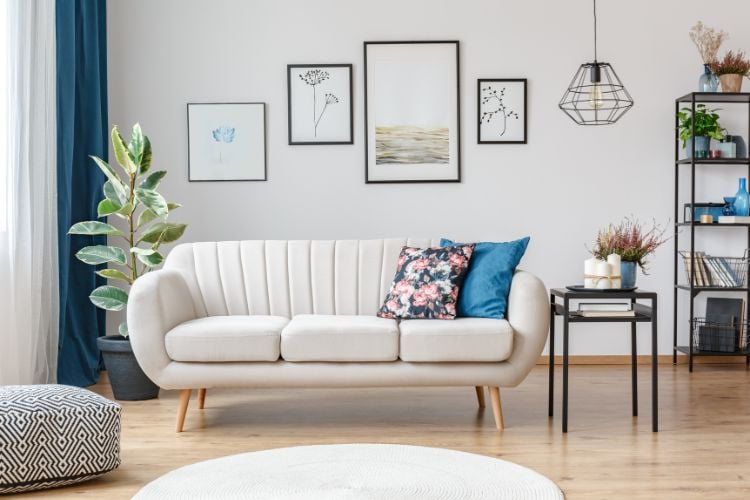 Sala com sofá off white arredondado, almofada floral, quadros fofos, tapete branco redondo e planta