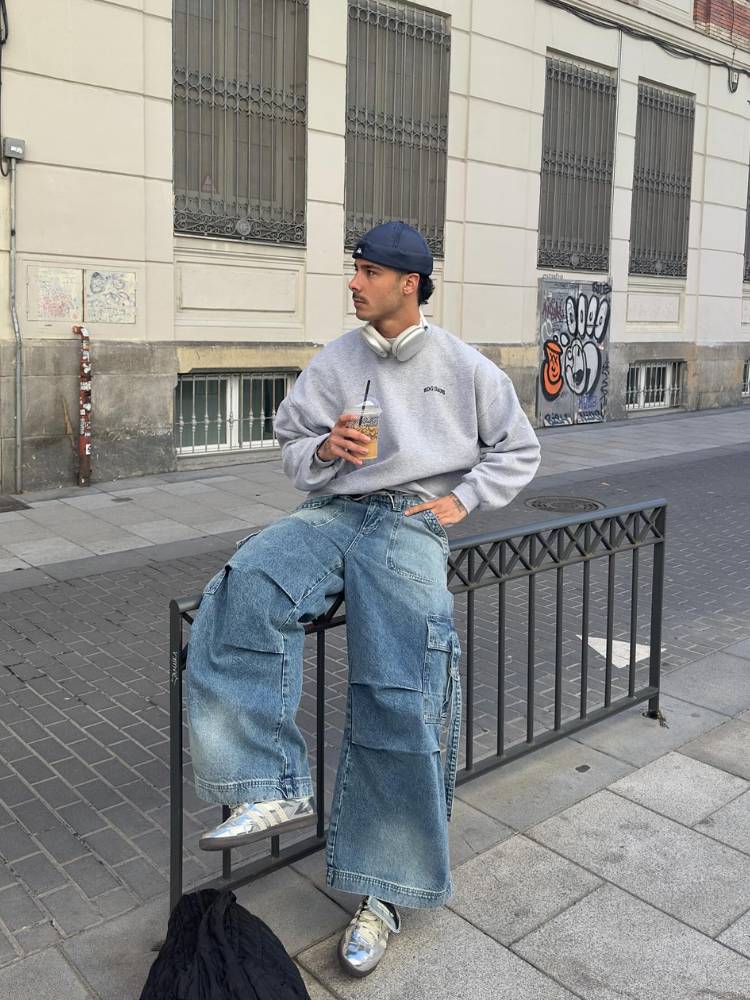 Homem de pele clara sentado em f=grade na rua usando calça jeans cargo, tênis, touca azul e suéter largo cinza