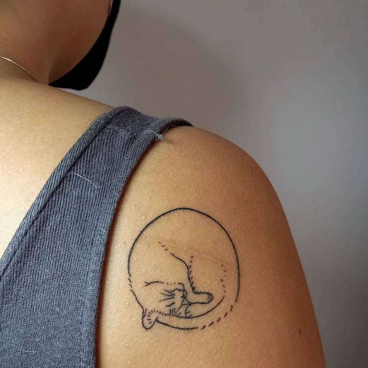 Mulher de pele morena com regata cinza e tatuagem na parte de trás do ombro, com o desenho redondo de um gato se lambendo