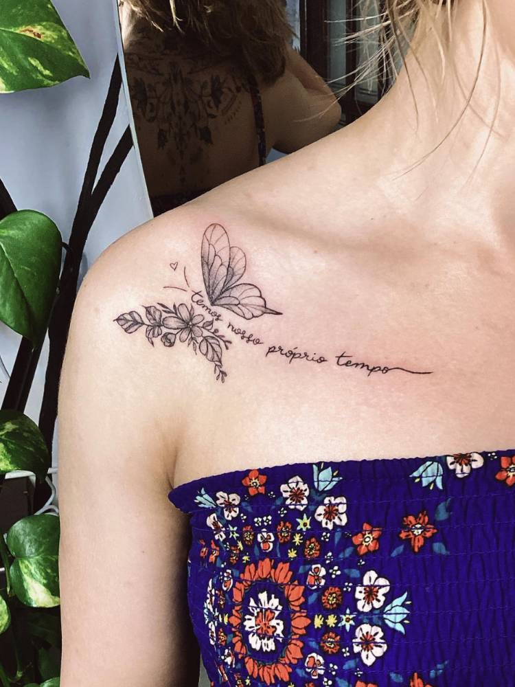Pessoa de pele clara com tatuagem de borboleta com uma asa de flores e frase "temos nosso próprio tempo"