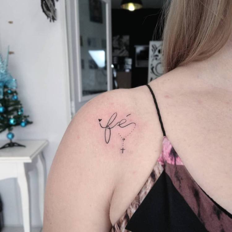 Mulher loira de pele clara usando regata preta e com tatuagem fine line escrito "fé"
