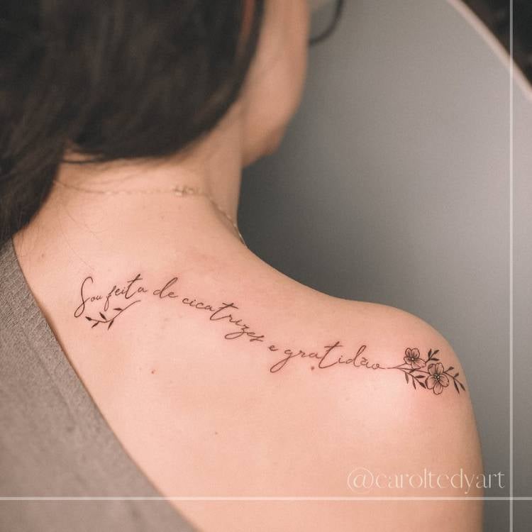 Mulher de pele clara de costas usando blusa cinza e tatuagem "Sou feita de cicatrizes e gratidão" com flor