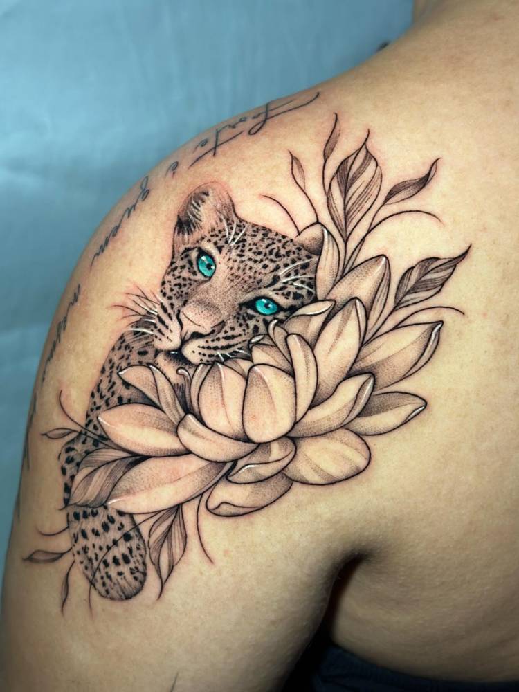 Mulher de pele clara com onça realista tatuada no ombro, com olhos azuis e abraçada com flores