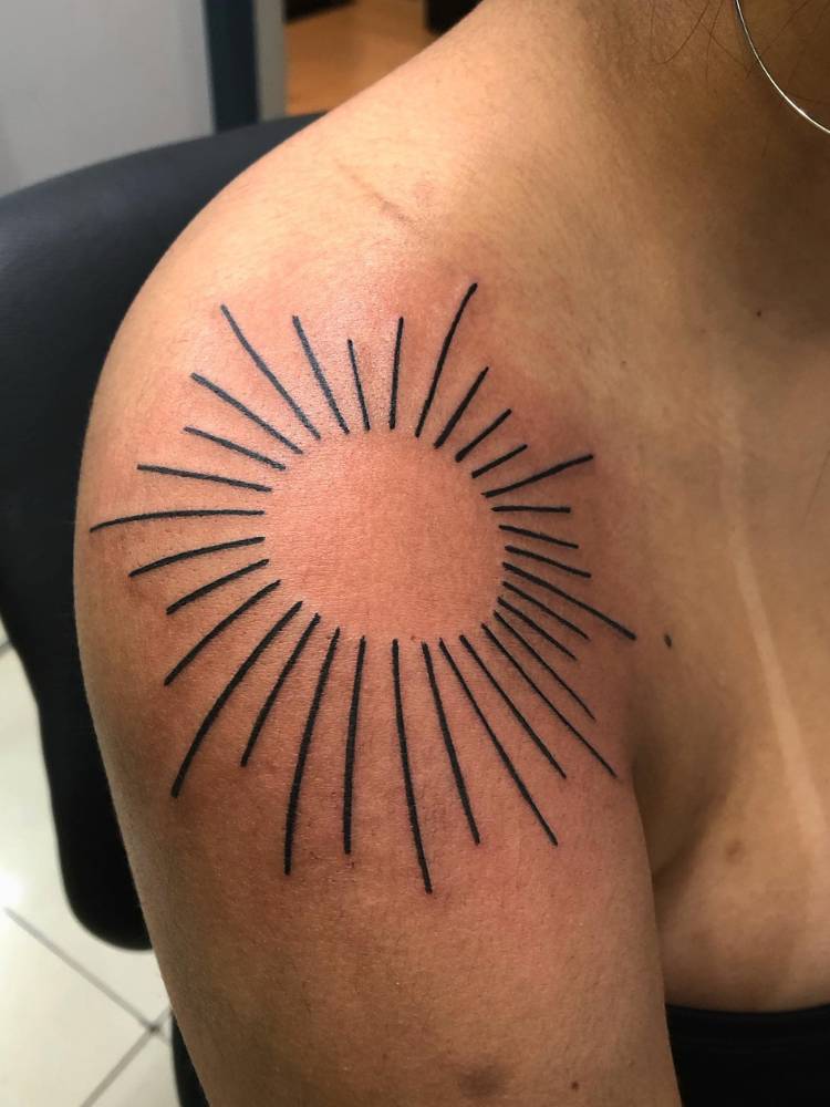 Mulher de pele morena com tatuagem de sol no ombro, apenas com os raios e círculo vazado, em traço preto grosso