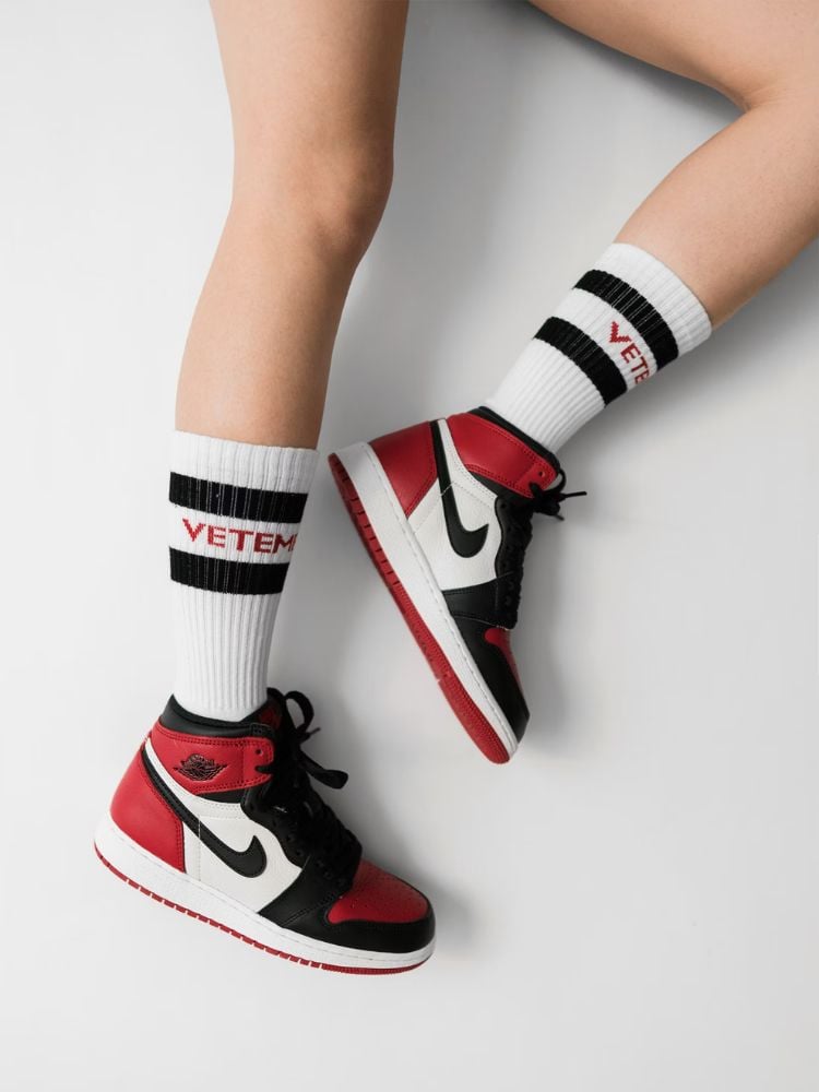 fundo branco com pernas de mulher usando meias brancas e tênis Nike vermelho, preto e branco nos pés