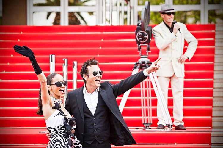 foto de casal de famosos usando roupas de gala e óculos de sol em tapetes vermelhos. A mulher usa vestido branco e preto com luvas brancas, enquanto o homem está de smoking preto