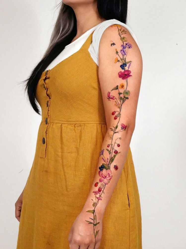 mulher morena usando blusa branca, vestido amarelo e tatuagem de flores coloridas ao longo de todo o braço