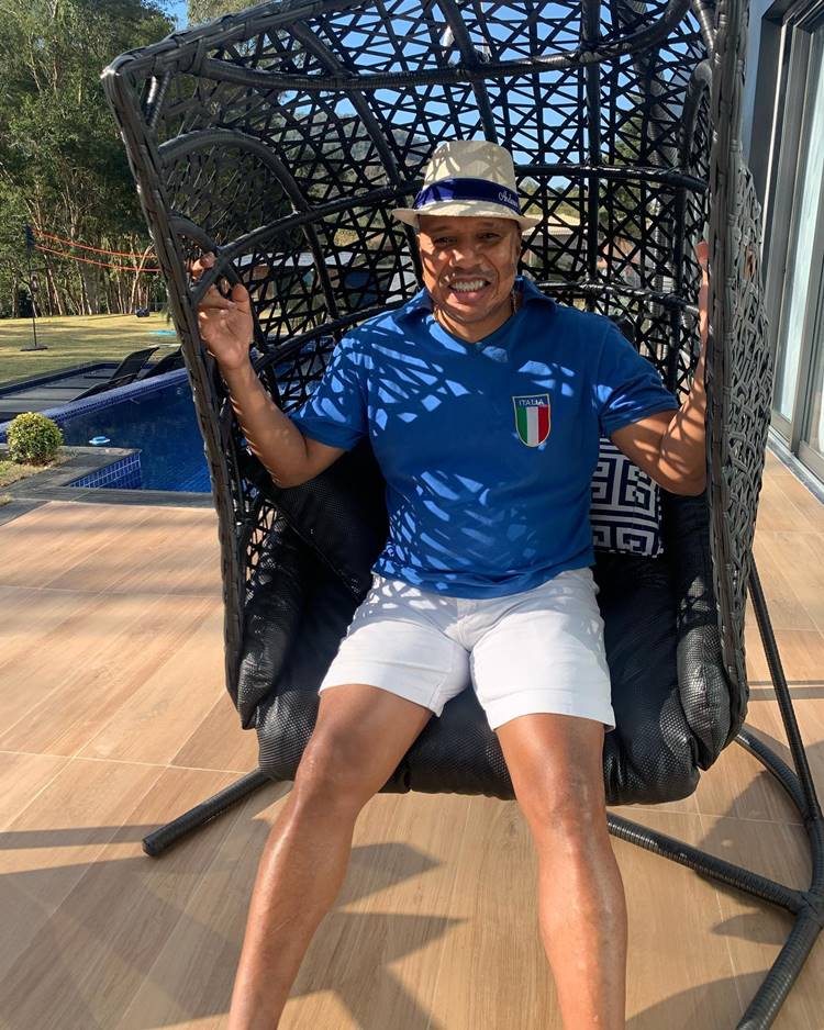 Cantor Anderson Leonardo com roupa azul, shorts branco e chapéu sentado em uma cadeira de balanço em uma área de piscina