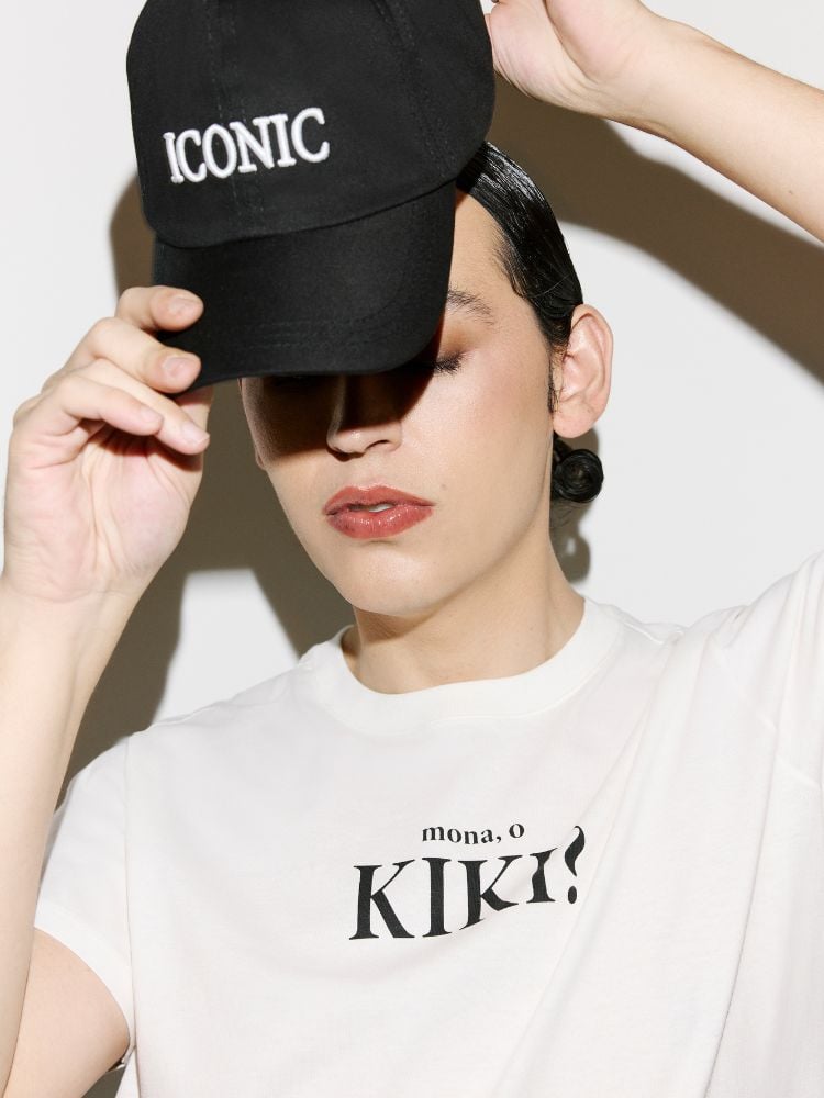 Gabb usando camiseta branca escrito "Mona, o kiki?" e colocando boné preto escrito "iconic"