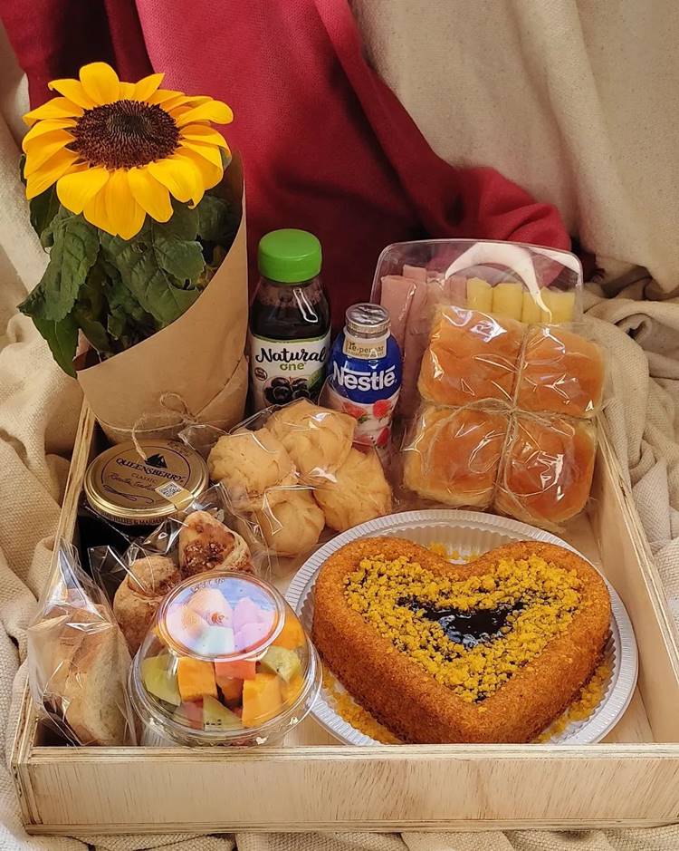Cesta de Dia das Mães com bolo de cenoura com chocolate, doces, pães, geleias, frios, e flor de girassol