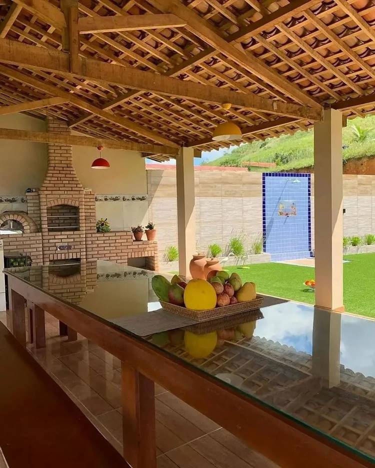 Cesta de frutas em mesa de madeira com tampo de vidro em uma cozinha com fogão à lenha e churrasqueira de tijolinhos